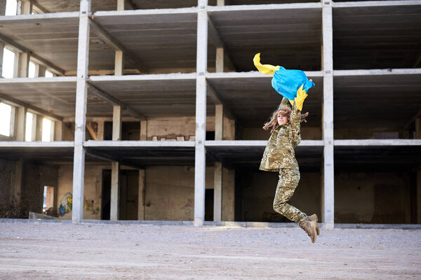 Молодая кудрявая блондинка, в украинской военной форме, держит украинский синий и желтый флаг, прыгает в воздух. Портрет женщины-солдата перед разрушенным заброшенным зданием
