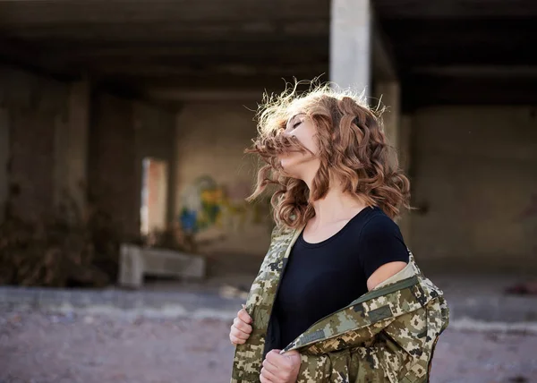 Young military woman, wearing ukrainian army military uniform and black t-shirt waving her curly blond hair in rim light. Three-quarter portrait of female soldier in front of ruined abandoned building