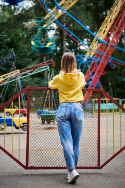 Sarışın, sarı kapüşonlu ve kot pantolonlu genç bir kadın yazın eğlence parkında vakit geçiriyor. Renkli dönme dolaba bakan güzel bir kızın tam boy arka portresi. Pazar keyfi.
