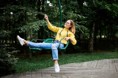 Sarı kapüşonlu ve mavi kotlu genç sarışın kadın, atlı karıncaya biniyor, gülümsüyor, yazın yeşil ağaçlarla parkta gülüyor. Eğlence parkında eğlence. Pazar boş vakti..