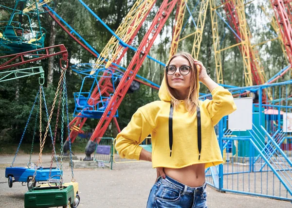 若いブロンドの女性は 黄色のパーカーと青いジーンズを着て 夏に遊園地で時間を過ごしています カラフルな観覧車の前でポーズをとって きれいな女の子の4分の3の肖像画 日曜日のレジャー — ストック写真