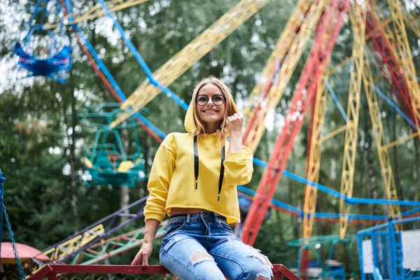 年轻的金发女人 穿着黄色的连帽衫和蓝色的牛仔裤 夏天呆在游乐园里 四分之三的女孩画像 坐在五彩斑斓的摩天轮前的栏杆上 面带微笑 — 图库照片