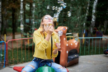 Sarı kapüşonlu, mavi kot pantolonlu ve gözlüklü genç bir kadın yeşil sarı ağaçlarla parkta eski bir atlıkarıncaya biniyor. Yetişkin bir kız, yazın parkta renkli sabun köpükleri yapıyor..