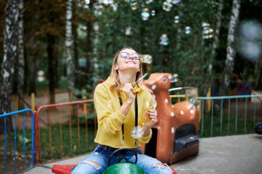 Sarı kapüşonlu, mavi kot pantolonlu ve gözlüklü genç bir kadın yeşil sarı ağaçlarla parkta eski bir atlıkarıncaya biniyor. Yetişkin bir kız, yazın parkta renkli sabun köpükleri yapıyor..