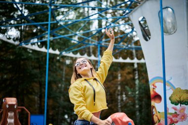 Sarı kapüşonlu, mavi kot pantolonlu ve gözlüklü genç bir kadın yeşil ağaçlı, renkli hayvanlarla dolu bir atlıkarıncaya biniyor. Yetişkin kız lunaparkta eğleniyor, gülüyor, gülümsüyor..