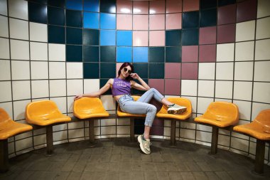 Bordo pembeli kısa saçlı, mor bluzlu, açık mavi kotlu ve siyah güneş gözlüklü, metro istasyonunda sarı koltuklarda oturan, yer altındaki yaratıcı kız portresi.