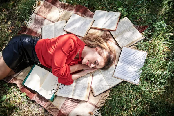 Young blond woman, wearing red shirt, lying sleeping on brown blanket. Creative flat-lay portrait, with person surrounded with many books. College student, tired on green grass after studying.