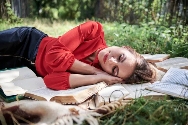 Young blond woman, wearing red shirt, lying sleeping on brown blanket. Creative close-up portrait, with person surrounded with many books. College student, tired on green grass after studying.