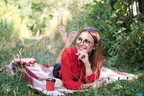 Young blond woman, wearing eyeglasses, red blouse and black skirt, lying on the blanket with books and coffee. Student, studying in the park. Leisure time at the college campus.