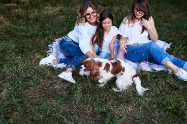 İki genç sarışın kadın ve bir esmer kız. Kot pantolon ve beyaz tişörtler giyiyorlar. Parkta yeşil bir battaniyenin üzerinde oturuyorlar. Yazın boş vakit..