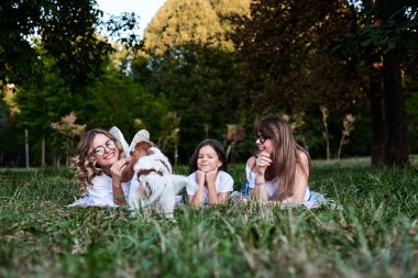 İki genç sarışın kadın ve beyaz tişörtler giyen esmer bir kız parkta yeşil çimenlerin üzerinde Meksika battaniyesine uzanmış, süvari kral Charles Spanel ile oynuyor, gülümsüyor, gülüyor. Boş zaman..