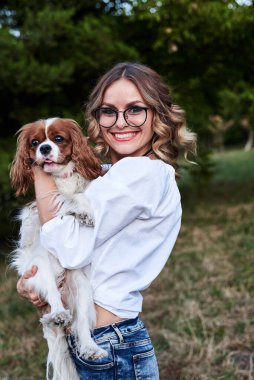 Kıvırcık saçlı, gözlüklü, beyaz tişörtlü, küçük beyaz ve kahverengi köpeği olan genç, güzel sarışın kadın yazın gülümsüyor. Şövalye Kral Charles Spaniel sahibiyle yürüyüşe çıktı..
