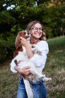 Kıvırcık saçlı, gözlüklü, beyaz tişörtlü, küçük beyaz ve kahverengi köpeği olan genç, güzel sarışın kadın yazın gülümsüyor. Şövalye Kral Charles Spaniel sahibiyle yürüyüşe çıktı..