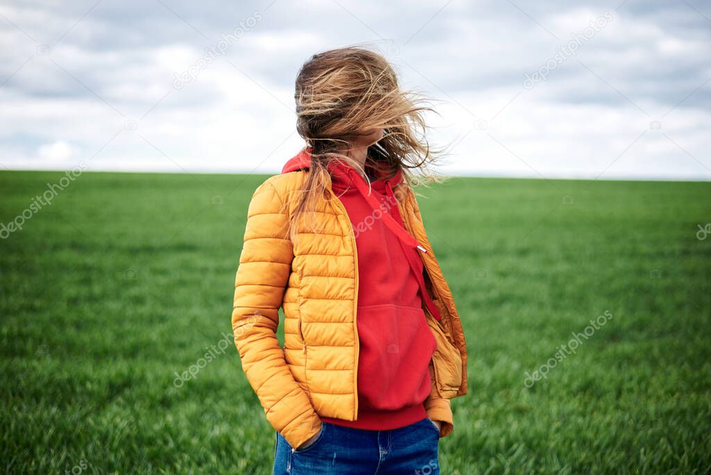 Young blond woman, wearing red hoody, yellow jacket and blue jeans, standing on green field with messy hair at windy weather in spring. Quarantine getaway in countryside. Stay home, isolate yourself.