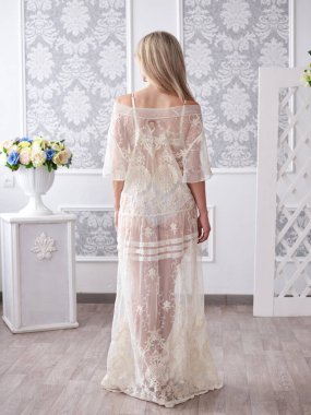 Uzun beyaz dantelli Peignoir giyen genç sarışın gelinin uzun boylu portresi, beyaz mobilyalarla aydınlık odada duruyor, düğün sabahı sırtı kameraya dönük,