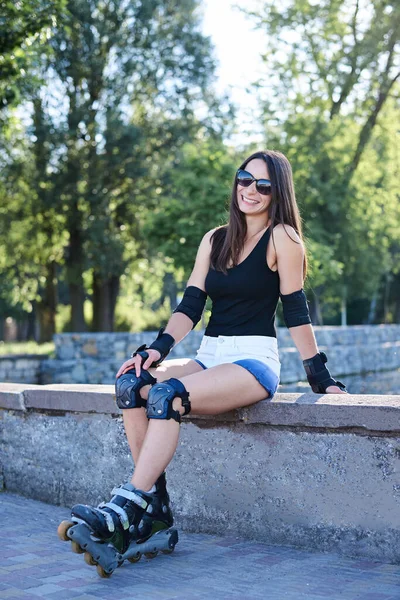 年轻漂亮的黑发女子 身穿轮滑刀片和安全设备 坐在混凝土平台上 骑完车后在公园里放松 轮滑运动健美女子的全长坐姿肖像 — 图库照片