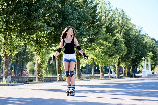 年轻美丽的黑发女子 骑着轮滑刀片在城市公园的早晨 适合运动的女孩 身穿黑色上衣和白色短裤 滑旱冰运动 苗条女运动员的全长肖像 — 图库照片