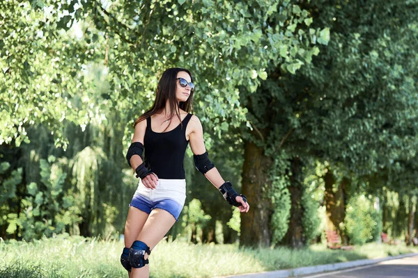 若いかなりブルネットの女性は 緑の木々と都市公園でローラーブレードに乗る 夏に黒の上と白のショートパンツ ローラースケートを着て スポーティな女の子をフィット スリムなスポーツ女性の3四半期の肖像画 — ストック写真