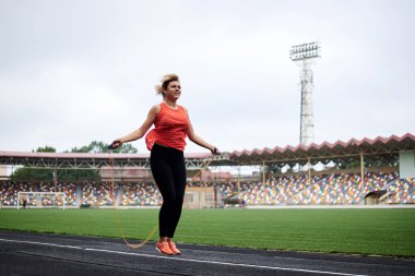 Sporcu kadının, stadyumda ip atlarken çekilmiş uzun bir resmi. Genç sarışın kadın, turuncu bluz ve siyah tayt giyiyor, yazın spor yapıyor..