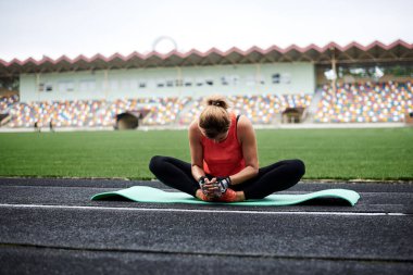 Turuncu bluz ve spor ayakkabı giyen, yeşil yoga minderinde oturan, kelebek yoga pozu vererek bacaklarını esneten sarışın genç bir kadın. Yazın stadyumda yoga eğitimi..