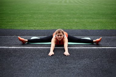 Genç sarışın kadın, yeşil çimlerle stadyumda sicim yapıyor. Güzel kız, turuncu bluz ve siyah tayt giyiyor, yazın bacaklarını ve vücudunu dışarıda açıyor. Fitness egzersiz eğitimi.