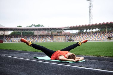 Genç sarışın kadın, yeşil yoga minderine uzanmış, düz bacaklarını sarı bilek ağırlıklarıyla kaldırıyor. Güzel sporcu kadın, turuncu bluz giyiyor, siyah tayt giyiyor, bacaklarına egzersiz yaptırıyor.