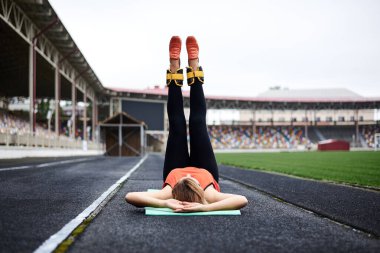 Genç sarışın kadın, yeşil yoga minderine uzanmış, düz bacaklarını sarı bilek ağırlıklarıyla kaldırıyor. Güzel sporcu kadın, turuncu bluz giyiyor, siyah tayt giyiyor, bacaklarına egzersiz yaptırıyor.