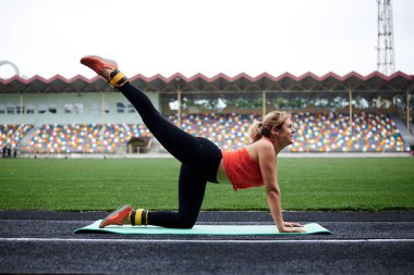 Genç sarışın kadın, bir diziyle yeşil yoga minderinin üzerinde duruyor, diğer bacağını vücudunun üzerine kaldırıyor, esniyor, sporcu kadın yaz aylarında yeşil çimlerle stadyumda spor yapıyor..