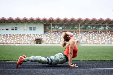 Turuncu bluz ve yeşil tayt giyen genç sarışın kadın, yaz aylarında yeşil çimlerle stadyumda kobra yoga pozu veriyor. Sağlıklı aktif yaşam tarzı konsepti. Yazın dışarıda spor yapıyor..