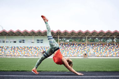 Turuncu bluz ve yeşil tayt giyen, aşağı bakan köpek yoga pozu veren, yaz aylarında yeşil çimlerle ayağını stadyuma kaldıran sarışın genç bir kadın. Sağlıklı yaşam tarzı kavramı. Fitness egzersizi