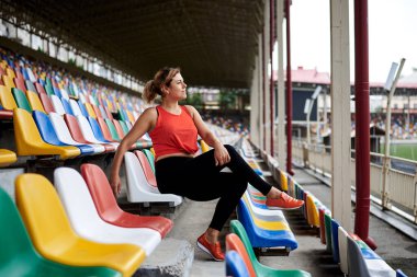 Genç sarışın kadın, turuncu bluz giyiyor, siyah tayt giyiyor, turuncu spor ayakkabılar, stadyumdaki renkli koltukta oturuyor, antrenmandan sonra bacaklarını tırabzanlara dayayıp dinleniyor. Boş Tribünde Sporcu Kadın.