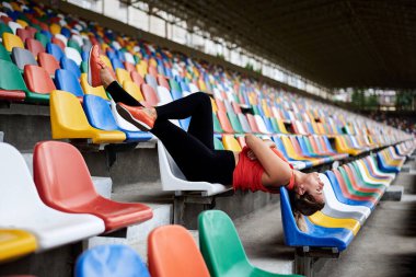 Turuncu bluz giyen, siyah taytlı, turuncu spor ayakkabılı, spor salonundaki renkli koltuklarda uzanan, antrenmandan sonra dinlenen genç sarışın kadın. Boş Tribünde Sporcu Kadın.