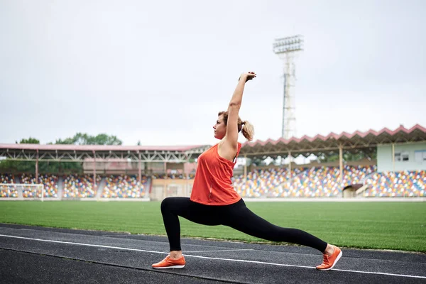 Young blond female athlete, wearing orange top and black leggings, doing lunges on a stadium with green grass in summer. Fit sporty woman is doing fitness exercises with motivation to loose weight.