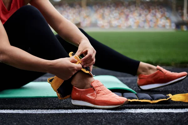 Close-up picture of woman\'s legs, showing process of putting on yellow ankle weights. Young woman, wearing black leggings and orange sneakers, is preparing herself for fitness training with weights.on
