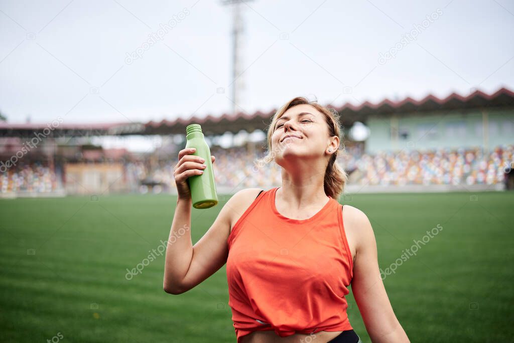 Young fit blond woman, wearing orange top and green leggings, drinking savoring water from light green bottle, smiling. Refresh during sport exercise on stadium with green grass. Athlete on training