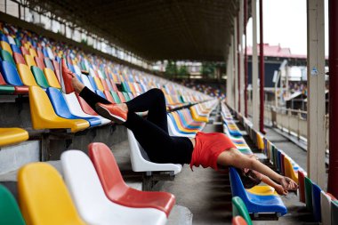 Turuncu bluz giyen, siyah taytlı, turuncu spor ayakkabılı, spor salonundaki renkli koltuklarda uzanan, antrenmandan sonra dinlenen genç sarışın kadın. Boş Tribünde Sporcu Kadın.