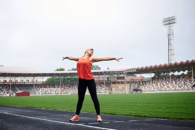 Sarışın bir kadının uzun bir portresi, yazın stadyumda esneme egzersizleri yapıyor, formda sporcu bir kadın, siyah tayt, turuncu bluz ve spor ayakkabılar giyiyor, antrenmandan önce kendini uyarıyor..