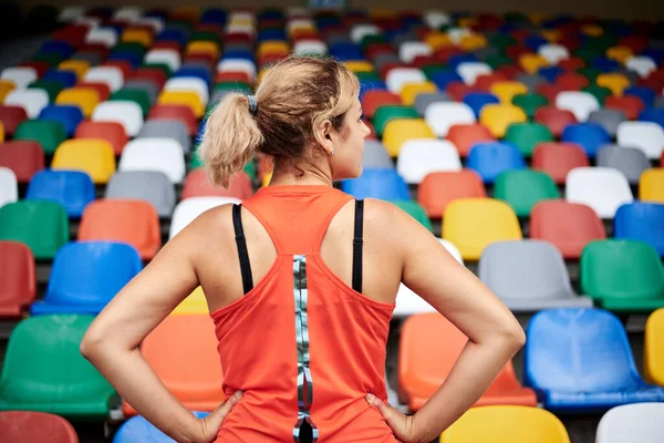 年轻的金发女子 身穿橙色上衣 背对着镜头 准备在空荡荡的体育场讲台上跑 座位五彩缤纷 夏季外出参加健身训练的女运动员 — 图库照片