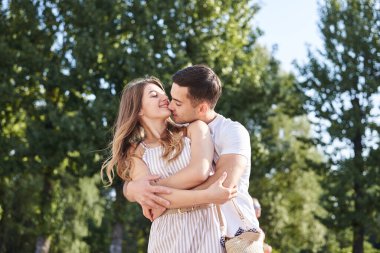 Yakışıklı esmer adam, beyaz tişört giyiyor, sarılıyor ve güzel sarışın kadını öpüyor. Genç aşıklar, yazın yeşil parkta kucaklaşıyorlar. Romantik bir ilişki. Sevgililer Günü.