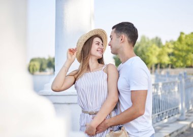 Yakışıklı bir adam, beyaz tişört giyiyor, mavi şort giyiyor, açık saman şapkalı ve çizgili yaz manzaralı güzel bir kadının arkasından sarılıyor. Genç çift birbirine aşık, sarılıyor, birbirlerine bakıyor, öpüşüyorlar..