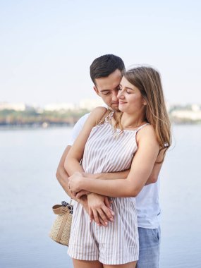 Yakışıklı esmer adam yazın Blue Lake 'in önünde güzel sarışın kadına arkadan sarılıyor. Genç çift birbirine aşık, sarılıyor, birbirine bakıyor, gülümsüyor, öpüşüyor..