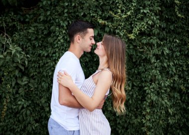 Genç çift, yeşil çalıların yanında birbirine sarılıyor. Güzel sarışın bir kadın, kısa çizgili bir şort giyiyor ve romantik bir randevuda beyaz tişörtlü ve mavi şortlu esmer bir adam. Romantik ilişki