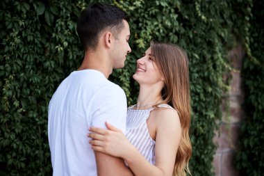 Genç çift, yeşil çalıların yanında birbirine sarılıyor. Güzel sarışın bir kadın, kısa çizgili bir şort giyiyor ve romantik bir randevuda beyaz tişörtlü ve mavi şortlu esmer bir adam. Romantik ilişki