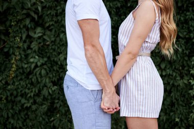 Beyaz tişört, mavi şort, çizgili yaz kıyafetleriyle sarılan bir kadın. Birbirini kucaklayan genç çiftlerin vücutları. Yeşil yaprak duvarının yanında romantik çift cesetlerinin yakın plan fotoğrafı.