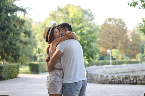 Young couple in love in park. Romantic date. Pretty woman, wearing stripy summer overall and straw hat is hugging and kissing attractive man in white t-shirt and light blue shorts. Love relationship