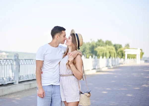 Handsome brunette man, wearing white t-shirt, blue shorts, is hugging pretty blond woman in light straw hat and stripy summer overall. Young couple in love, embracing, looking at each other, smiling.