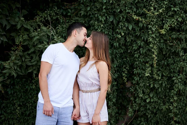 Young couple in love hugging kissing near green bushes trees wall. Pretty blond woman, wearing stripy short overall and brunette man in white t-shirt and blue shorts on romantic date. Relationship