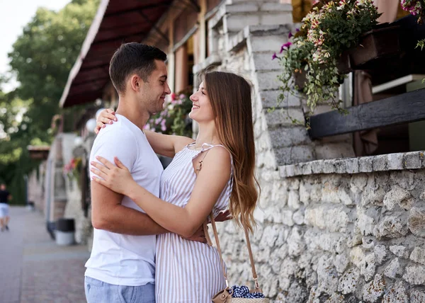 Young couple in love hugging kissing near stone wall in resort town. Pretty blond woman, wearing stripy short overall and brunette man in white t-shirt and blue shorts on romantic date. Relationship