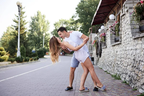 Young couple in love hugging kissing near stone wall in resort town. Pretty blond woman, wearing stripy short overall and brunette man in white t-shirt and blue shorts on romantic date. Relationship