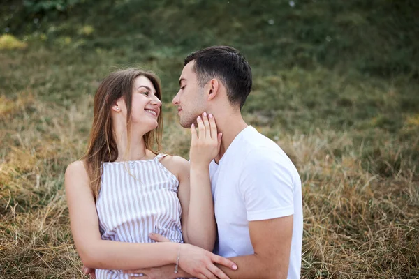 年轻夫妇坐在翠绿的黄草草坪上拥抱亲吻 在浪漫的约会中 金发女人总体上穿着条纹短裙 黑发男人穿着白色T恤蓝色短裤 — 图库照片